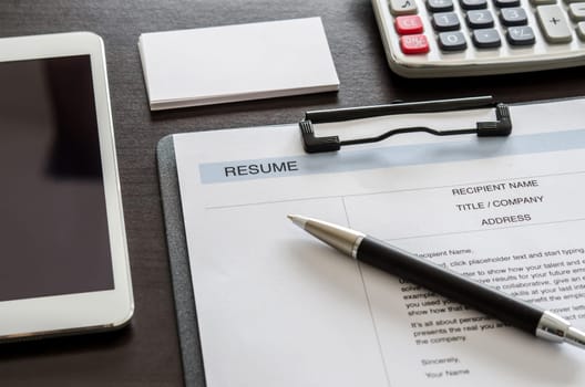 Close-up resume form, pen, digital tablet, calculator and business card on wooden desk.