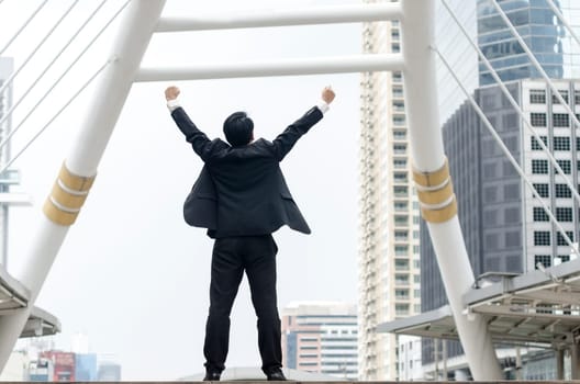 Success concept. Backside of asian businessman hands up with happy moment and modern building background.