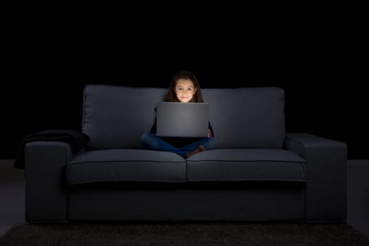 Happy little girl sitting on a couch and working a laptop