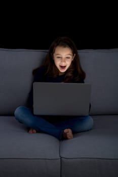 Happy little girl sitting on a couch and working a laptop