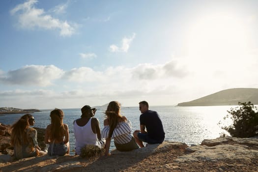 Rear View Of Friends Sitting On Cliff Watching Sunset