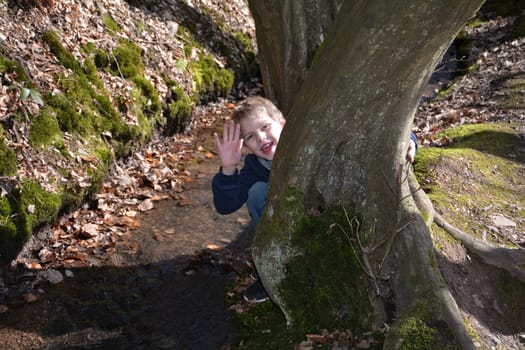 Little boy by the stream,kneels behind a tree and waves forward laughing