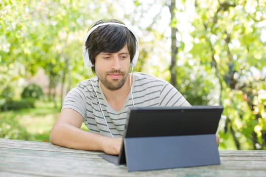 young man working with a tablet pc listening music with headphones on a wooden table, outdoor