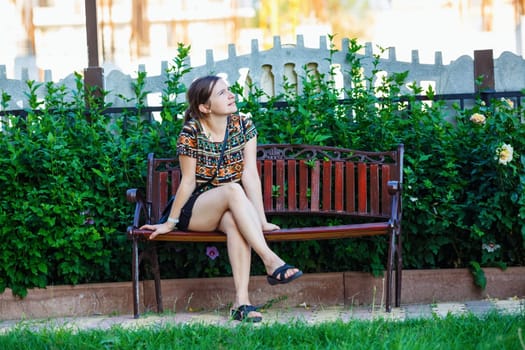 Pretty young woman sitting on a bench in the park on background of bright green foliage. Selective focus on girl.