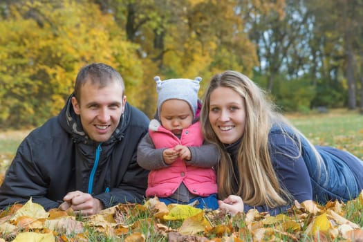 Smiling young parents with their baby daughter are lying on the ground and looking at the camera in a beautiful autumn park