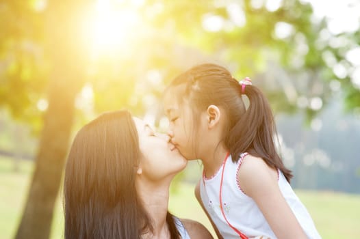 Lifestyle portrait mom and daughter kissing in happiness at the outside in the park. Family outdoor fun, morning with sun flare.