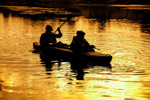 Silhouette of special forces men paddling army kayak. Boat moving calmly across the river, diversionary mission, sunset dusk