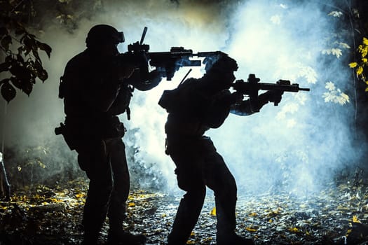 Black silhouettes of pair of soldiers in the smoke haze moving in battle operation. Back light