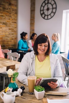 Mature woman is sitting in a cafe using a digital tablet while enjoying a cup of coffee.
