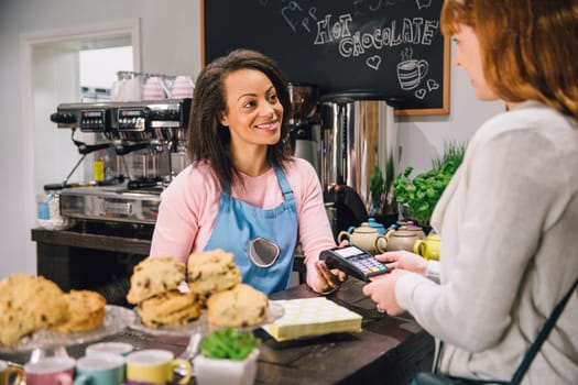 Woman is making a card payment at the counter in a cafe. The woman behind the counter is holding the card machine and smiling to the customer.