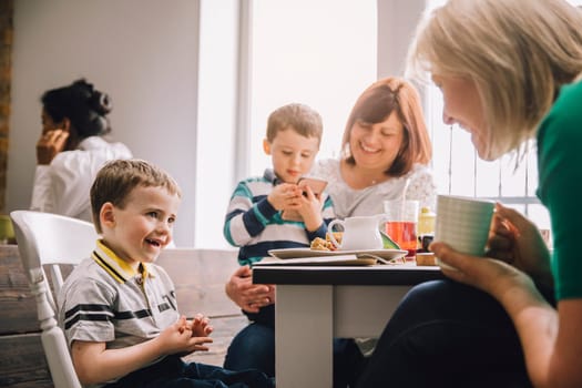 Litle boy is talking to his mothers friend in a cafe. The mother can be seen out of focus in the background with her other son on her lap, playing with her smartphone.