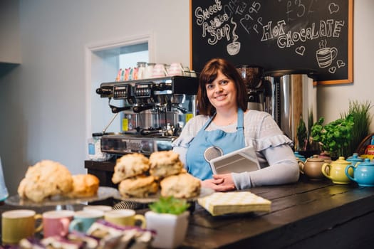 Friendly waitress is standing behind the counter in the cafe she works in and is smiling for the camera with a digital tablet.