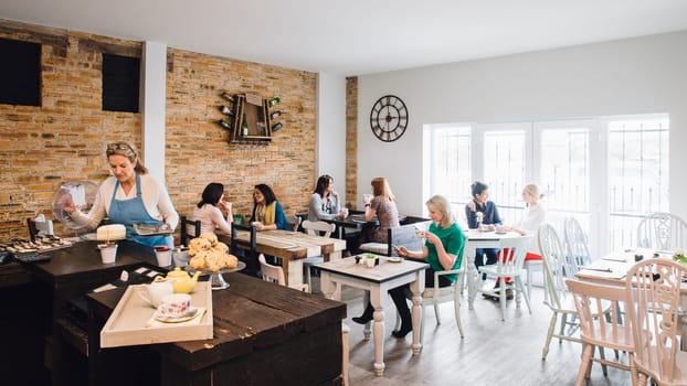 Room shot of a busy cafe full of women socialising. There is a mature waitress serving cake from a counter.