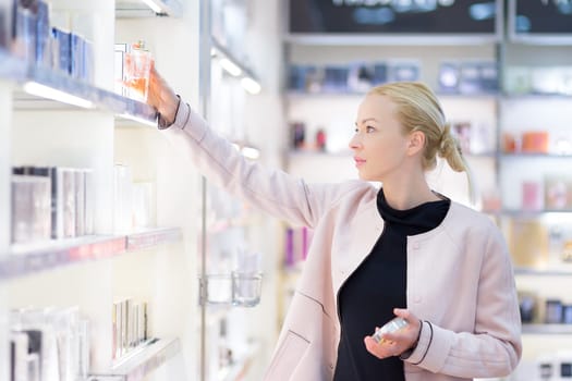 Elegant blond young woman choosing perfume in retail store. Beautiful blond lady testing and buying cosmetics in a beauty store.