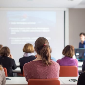 Female speaker giving presentation in lecture hall at university workshop. Rear view of unrecognized participants listening to lecture and making notes. Scientific conference event.