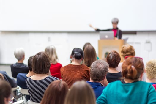 Business and entrepreneurship symposium. Female speaker giving a talk at business meeting. Audience in conference hall. Rear view of unrecognized participant in audience. Copy space on whitescreen.