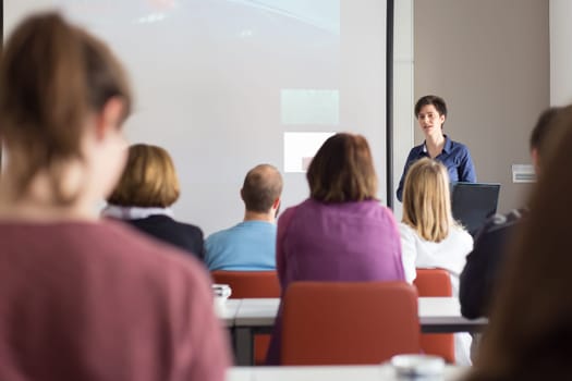 Female speaker giving presentation in lecture hall at university workshop . Participants listening to lecture and making notes. Scientific conference event.
