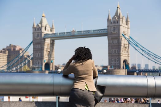 Businesswoman talking on mobile phone outdoor, looking at Tower Bridge in London city, UK.