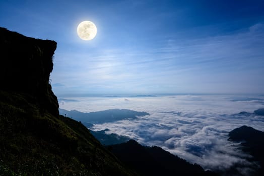 Beautiful landscape nature, Full moon with bright white light over the clouds and fog in the dark blue sky at night on the peak mountain in winter at Phu Chi Fa Forest Park, Chiang Rai Province, Thailand