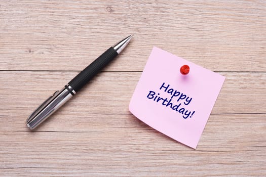 Concept Happy Birthday on pink sticky note with ballpoint pen on wood