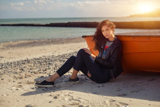A girl sideways sitting near a red boat on the beach by the sea with beautiful sunset on the background. Woman in jeans, pullover and jacket on the sand