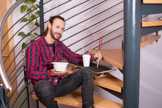 young man in checkered shirt sitting on stairs and eating asian food