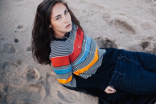 Young girl on the beach lying on the sand. She wearing in jeans and colored sweater. Woman with long hair in a colored line pullover on the beach. A woman enjoys life.