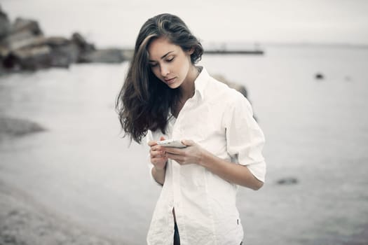 Portrait of teenager girl walking on the beach is checking online the mobile phone waiting for a message. She wear on the white shirl.