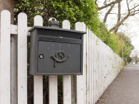 an interesting and unique black post box outside locked with an icon and attached to a white fence on a path and with hedge in spring clear delivery home