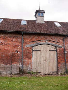 An Old Set of Wooden Doors on the Outside of a Building with red Bricks and A Chimney and Roof, big, brown and closed doors, aged, interesting, powerful, shot from the grass at the floor