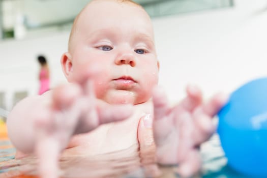 Newborn baby in pool at infant swimming lesson reaching for water ball