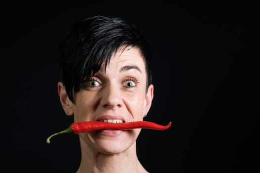 Head portrait of a mid-aged gay woman with a red chilli pod between her teeth in front of a black background looking into the camera.