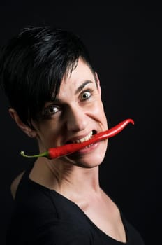 Head and shoulder portrait of a mid-aged gay woman with a red chilli pod between her teeth looking into the camera in front of a black bottom.