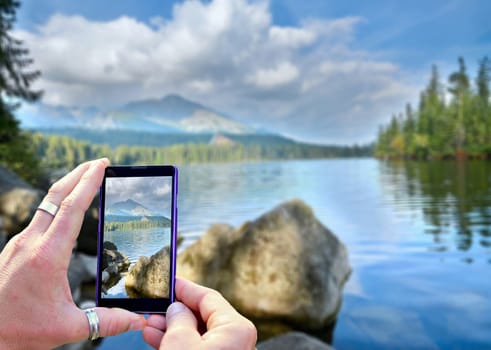 View over the mobile phone display during taking a picture of landscape in nature. Holding the mobile phone in hands and taking a photo. Focused on mobile phone screen.