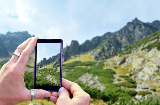 View over the mobile phone display during taking a picture of landscape in nature. Holding the mobile phone in hands and taking a photo. Focused on mobile phone screen.