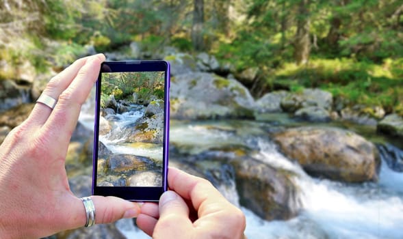 View over the mobile phone display during taking a picture of landscape in nature. Holding the mobile phone in hands and taking a photo. Focused on mobile phone screen.