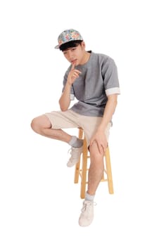 A young Korean teenager sitting on a chair in shorts and a cap with.his finger on his chin, isolated for white background..