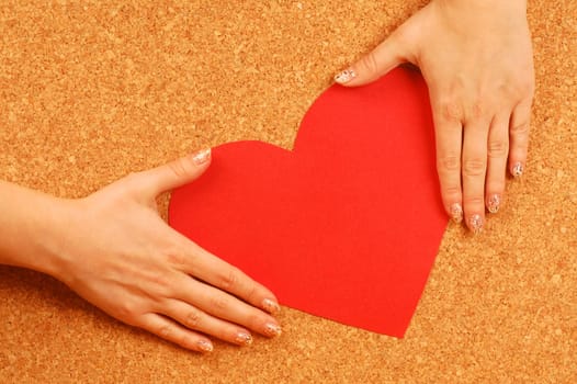 Female hands hold red heart from paper on cork background.