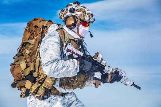 Army serviceman in winter camo somewhere in the Arctic. He wears chest rig and huge backpack, suffers from the extreme cold and strong wind, but endures while mission continues. Blue sky background