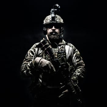Bearded soldier in Combat Uniforms with weapon, plate carrier and combat helmet are on. Studio shot, dark background. Ruthless and fearless, he brings death to enemies, defending his country
