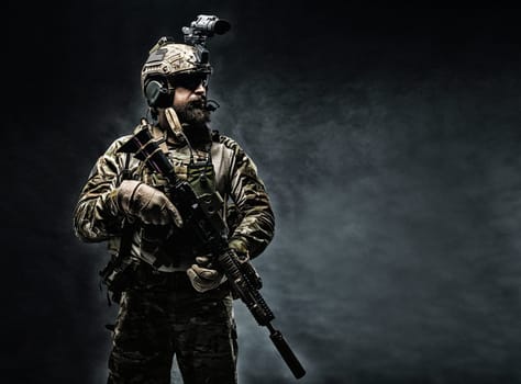 Bearded soldier in Combat Uniforms with weapon, plate carrier and combat helmet are on. Studio shot, dark background