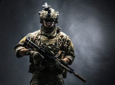Army soldier in Combat Uniforms with assault rifle, plate carrier and combat helmet are on, Shemagh Kufiya scarf on his neck. Studio shot, dark background