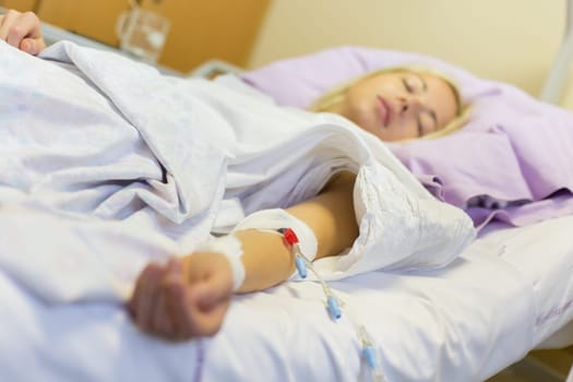 Bedridden female patient lying in hospital bed, receive infusion while recovering after surgery.