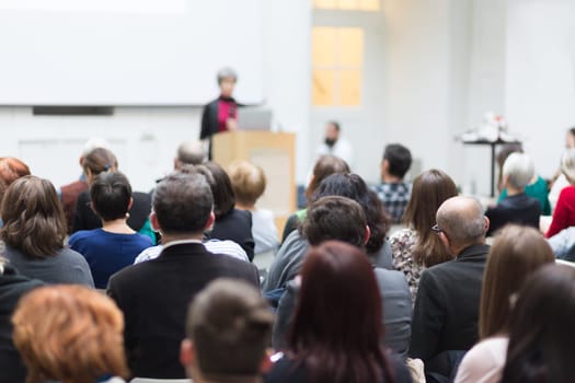 Business and entrepreneurship symposium. Female speaker giving a talk at business meeting. Audience in conference hall. Rear view of unrecognized participant in audience.