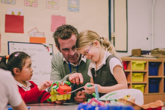 Primary school teacher is helping two of his students with a STEM project. They are building something using recycled items and crafts equipment.