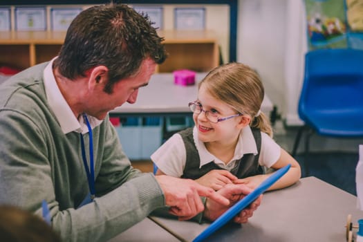 Male teacher is using a digital tablet with his student in a classroom lesson. He is talking to her and pointing to something on the screen of the tablet.