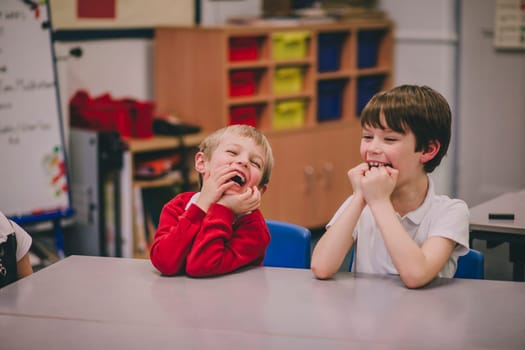 Two little boys are at school, sitting at a table in their classroom. They are both hysterically laughing at something.