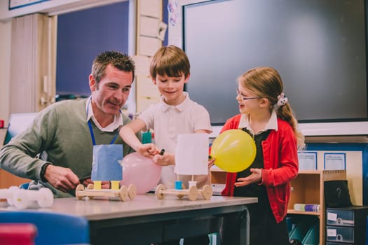 Primary school teacher is helping two of his students with a STEM project. They are building something using recycled items and crafts equipment.