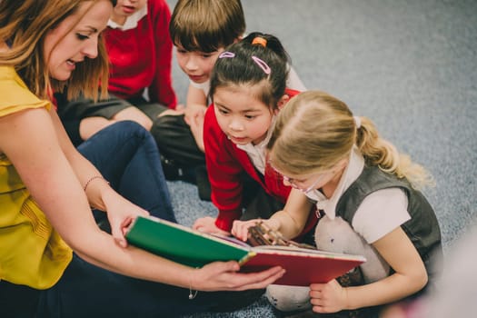 Teacher is sitting in the classroom with her primary school students, reading a story to them.