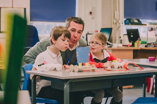 Teacher is sitting with two of his primary school students in the classroom, helping them test their physics project. They are creating air by blowing with their mouths, to move their toy vehicles.
